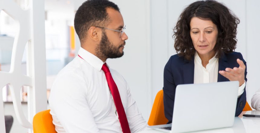 Project leader explaining job specific to newcomer. Business man and woman sitting at conference table with open laptop, talking and gesturing. Expertise concept